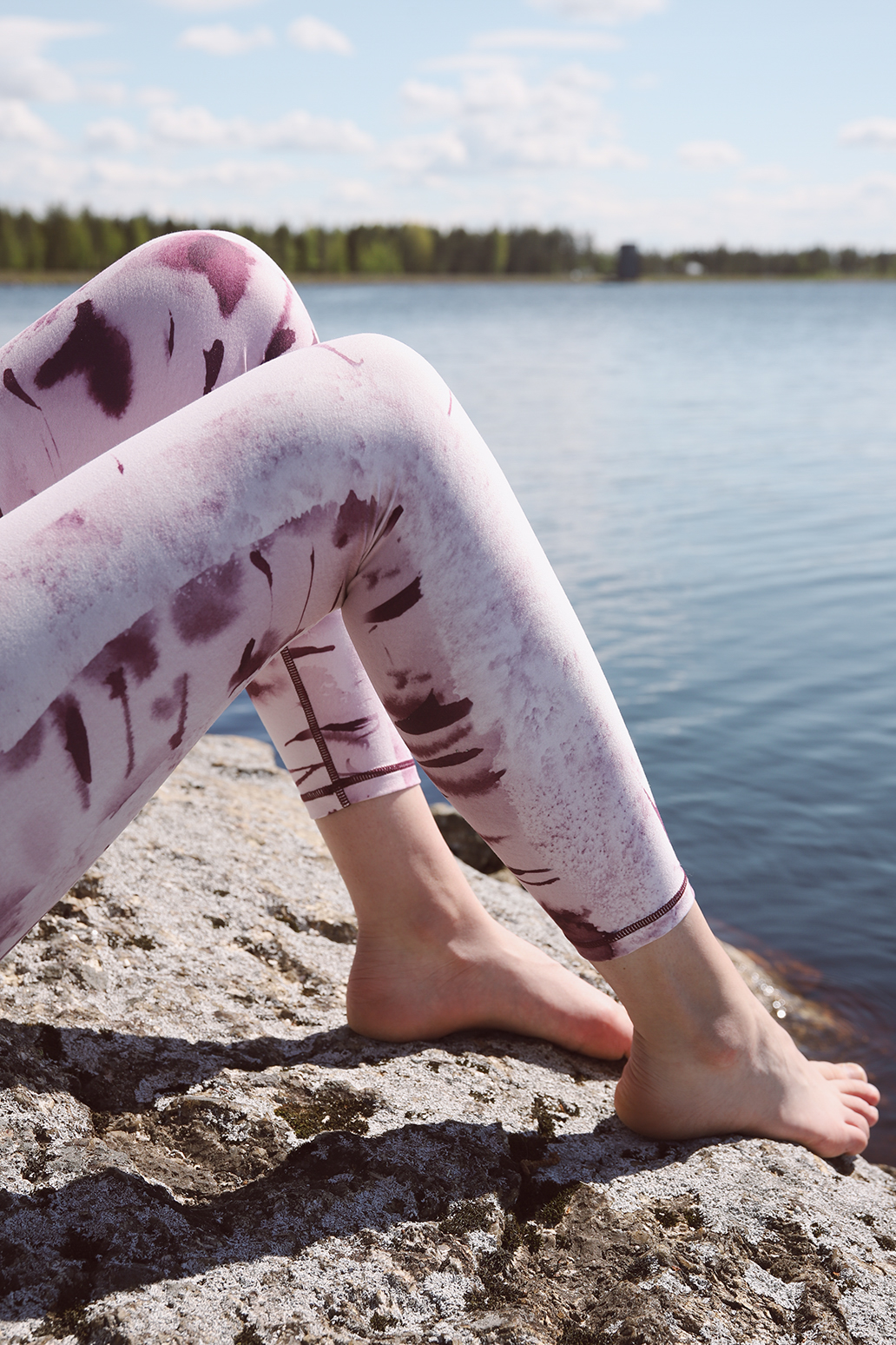 Close-up of crossed legs and bare feet wearing Silver birch - Soahki lily leggings with maroon tie-dye print on a rocky lakeshore