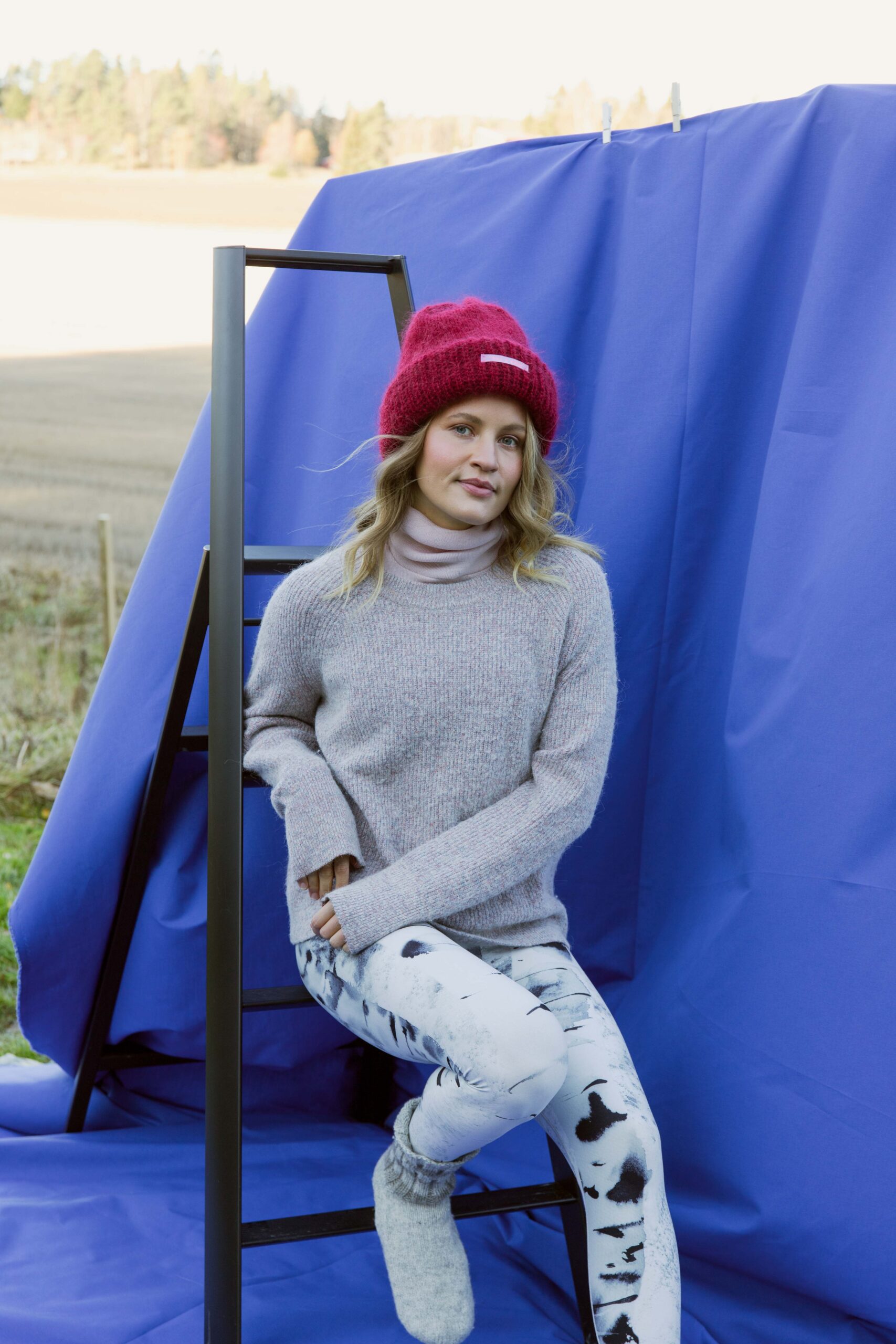 Seated model wearing Silver birch - Soahki black print leggings with knit sweater, red beanie and grey socks, studio backdrop outdoors