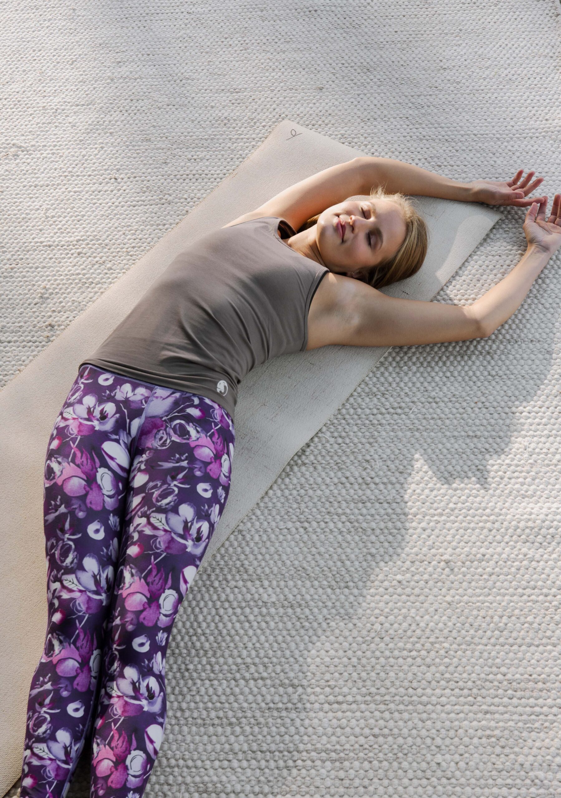 Woman lying on a yoga mat wearing Organic Shanti taupe tank top with Snowbed willow - Gironlasta lily purple floral leggings, arms extended overhead